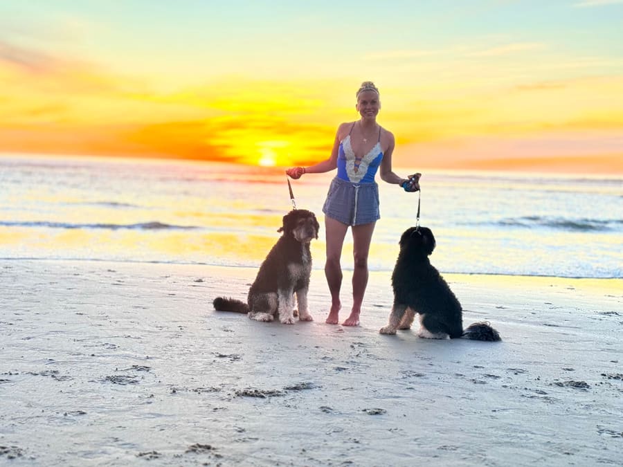 Elisabeth on the beach at sunset holding the leashes of two doodles sitting in the sand