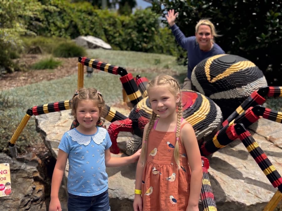 Two girls smiling in front of a giant LEGO spider while Elisabeth waves from behind it